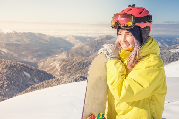 A smiling snowboarder stands at top of snow covered mountain holding snowboard