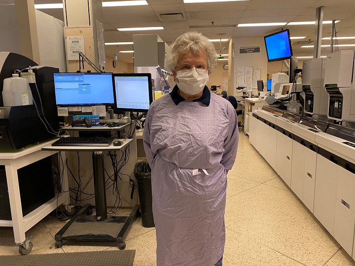 Betty Chapelski in face mask and lab coat, standing in lab next to computer monitors